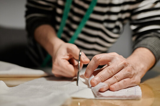 Cropped unrecognizable concentrated female designer in casual clothes standing near table and cutting fabric with scissors while working at home