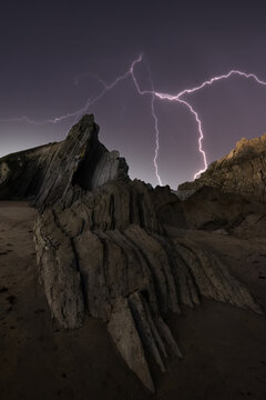 From below rough dramatic landscape of rocky cliff under dark stormy sky with bright lightnings at night