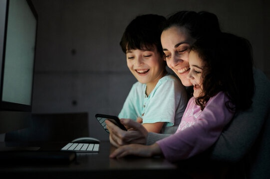 Happy young mother with little son and daughter holding smartphone while sitting together at table with computer and watching funny video in dark room at home