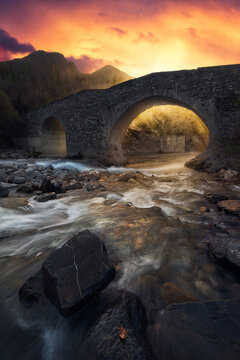 Amazing scenery with ancient stone bridge crossing shallow river against majestic mountains and colorful sunset sky in autumn evening in countryside