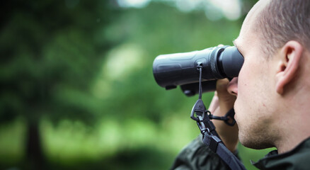 Panorama a young hunter in the forest looks into the binoculars. He is trying to find some wild animals.