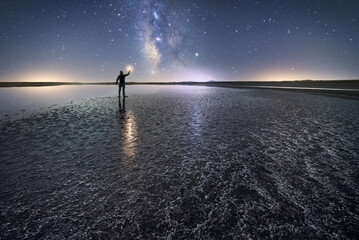 Back view of anonymous man standing and holding torch on empty road among calm water and reaching out to star under colorful nigh sky with milky way on background