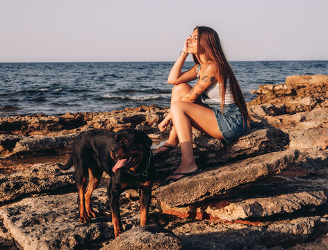 Side view of trendy tattooed woman looking away while sitting on stone with Rottweiler dog while spending time on rocky seashore