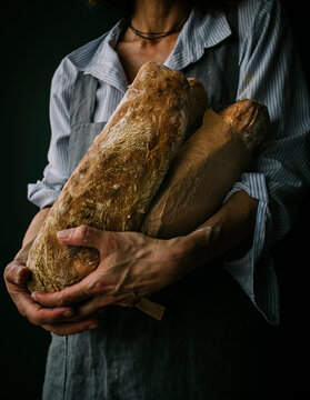 Cropped unrecognizable woman in apron holding ciabatta bread while standing on dark background