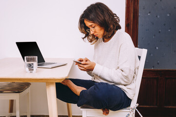 Side view of focused young female freelancer in casual clothes checking messages on smartphone while sitting at table with laptop and working on project at home