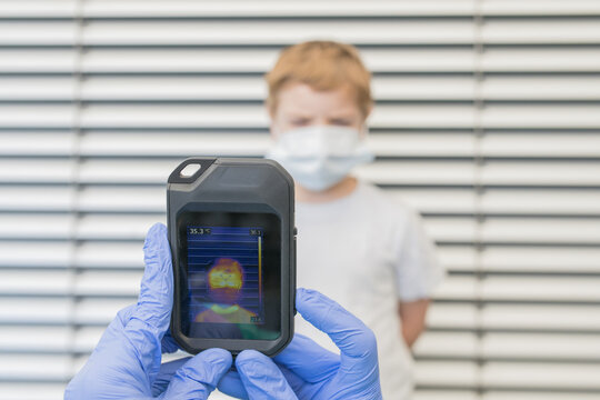 Unrecognizable medic in protective gloves and uniform using thermal imaging camera for checking temperature of kid on street during coronavirus outbreak