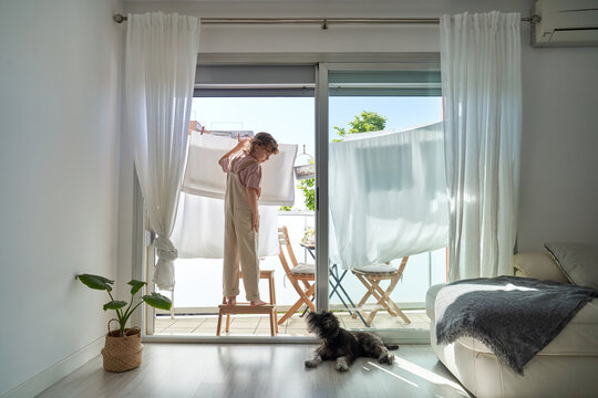 Back view of kid in casual clothes standing on chair on balcony and hanging wet linen on rope with dog lying down on the floor at home