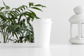 Coffee paper cup mockup with a candle holder and a palm plant on a white table.