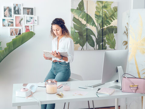 Beautiful female in elegant outfit cheerfully smiling and reading notes in notepad while leaning on table in stylish office