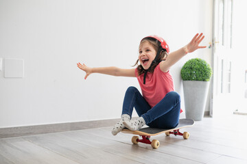 Little kid in red spotted helmet and casual clothes and dreaming about outdoors amusement while sitting alone on longboard in corridor of light modern apartment staying at home