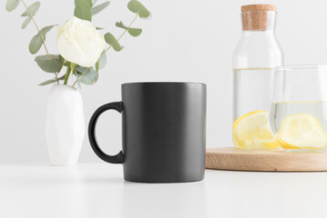 Black mug mockup with flowers in a vase, glass and a bottle on a white table.