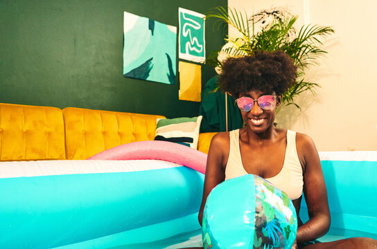 Cheerful ethnic female in bikini and sunglasses sitting in inflatable pool with ball while relaxing during party at home
