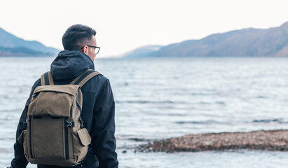Back view of male hiker in warm jacket with backpack sitting on wooden bench near sea and enjoying marine scenery with rocky coast during travel in Scotland