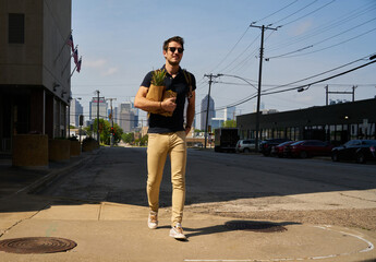 Handsome male in casual outfit and trendy sunglasses walking with grocery bags after shopping while looking away and enjoying sunny weather