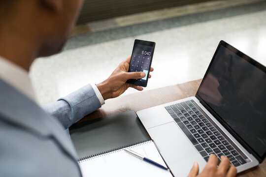 Crop man using laptop and doing calculation on smartphone sitting at table with notepad.