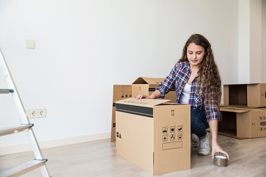 Cute female sitting near carton boxes and ladder using duct tape in empty apartment