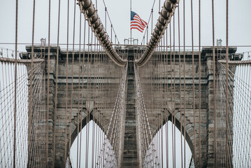 Low angle of stone towers with pointed arches of suspension Brooklyn bridge with American flag on cloudy day