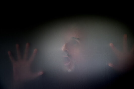 Unidentifiable male with mustache gray beard wide eyed open mouth screaming face hands pressed against veil or screen, showing anguish fear depression mental health psychological suffering copy space 