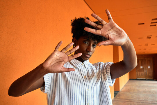 Adult African American female with eyes closed trying to stop negative impact while standing with arms raised against blurred vivid orange interior in corridor of modern building