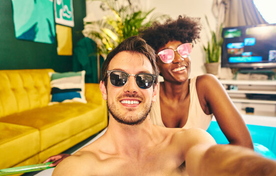 Cheerful multiracial couple wearing sunglasses sitting in inflatable pool and taking selfie while enjoying weekend