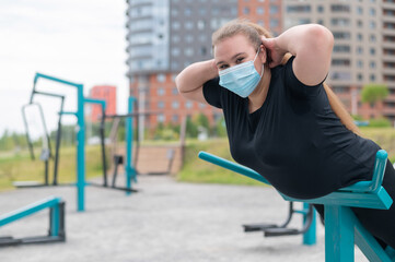 Overweight young woman in a protective mask plays sports on a playground. The girl does exercises for back muscles on the simulator outdoors.