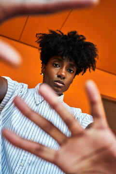 Adult African American female with eyes closed trying to stop negative impact while standing with arms raised against blurred vivid orange interior in corridor of modern building