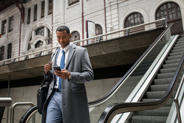 Handsome black businessman in coat and with phone walking down moving stairs while commuting.