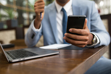 Crop man using laptop and doing calculation on smartphone sitting at table with notepad.
