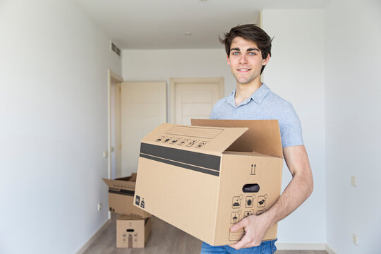 Handsome young male holding carton box in empty room looking at camera and smiling