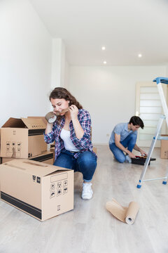 Young female cutting duct tape with teeth sitting near carton boxes and male sitting near black box in empty apartment
