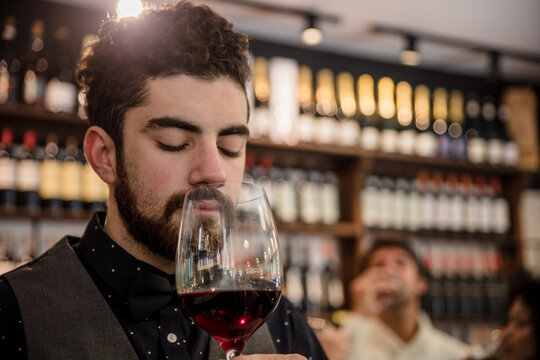 Bearded sommelier in elegant outfit keeping eyes closed and smelling red wine in glass.