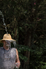 Older man gray beard goatee in sleeveless t-shirt with tattered straw hat & glasses, filling cup from heavy stream of water coming from above, country life, cooling down with water on a hot day