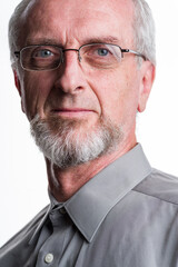 Man in his 50's or 60's with gray grey hair and beard or goatee and button-up gray shirt looking at camera with eye-glasses with confident but tired look in eyes, close-up portrait  white background