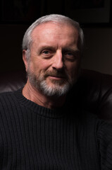 Obraz premium Middle aged man with gray hair and beard sitting comfortably in high backed chair with slight welcoming amused friendly smile with dark sweater and background