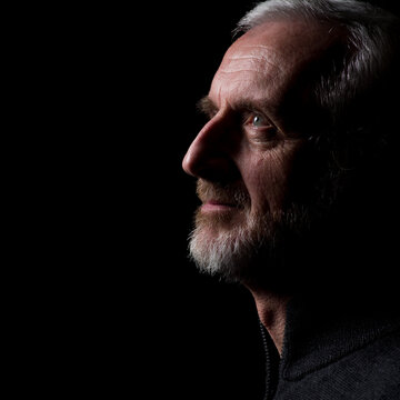 Middle aged man with gray hair and beard,  close up profile with hard shadows on black background with eyes open with face and gaze slightly upward in thought, prayer, meditation or contemplation
