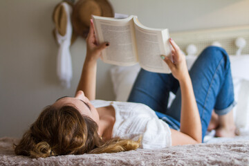 Chica joven tumbada boca arriba en la cama de su habitación leyendo un libro - obrazy, fototapety, plakaty