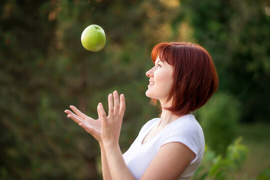 Beautiful Woman With Red Hair Catching Big Green Apple Falling From Above Outdoors. Cheerful Woman Trying To Catch Apple In Garden. Careful Planning, Success And Achieving Goals Concept