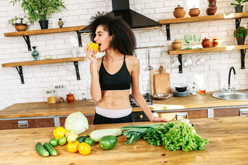 Eat healthy foods. Athletic african american girl in sportswear in the kitchen bites fresh pepper, vegetables and greens on the table
