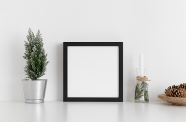 Black square frame mockup with a cypress tree, candles and pine cones on a white table. Christmas decoration.
