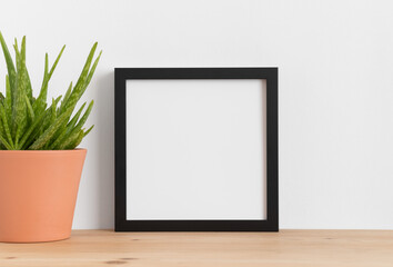 Black square frame mockup with a aloe vera in a ceramic pot on a wooden table.