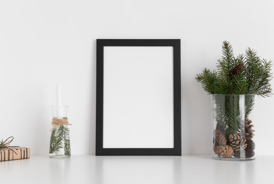 Black frame mockup with a bouquet of fir, candle and a gift on a white table. Christmas decoration.