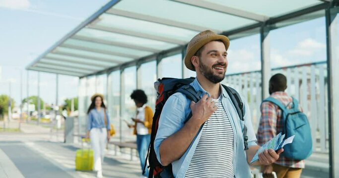 Caucasian young handsome man traveller in hat walking outdoors and carrying backpack. Good-looking happy male tourist with map strolling at bus and tram stop and smiling.