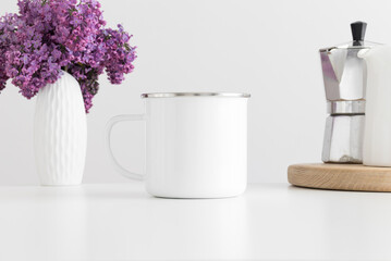 Enamel mug mockup with a moka pot and a bouquet of lilac in a vase on a white table.