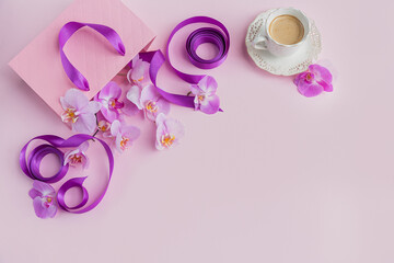 Flower overhead composition on light pink background top view. Cup of coffee, pink gift bag with purple ribbons and pink orchid flowers.