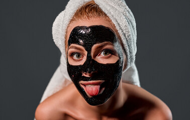 Portrait of a young attractive woman with a towel on her head with a black cleansing mask on her face shows tongue isolated on a gray background.