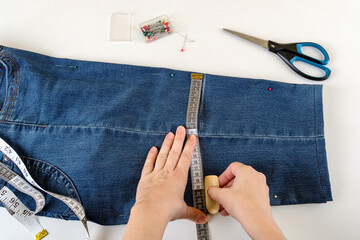 Caucasian woman hands drawing a cut line on a folded in half blue denim capris on a white table. Shorten the jeans with scissors and sewing pin. DIY summer clothes.