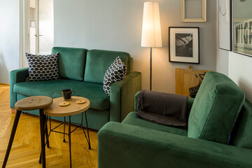 wnętrz - ciemnozielona sofa i fotel, obok lampa, stolik i taboret, na podłodze parkiet drewniany