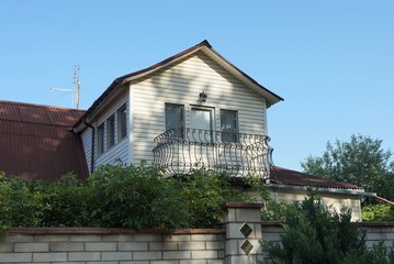 one brown iron balcony in the white attic of a private house with a brown roof overgrown with green vegetation against a blue sky