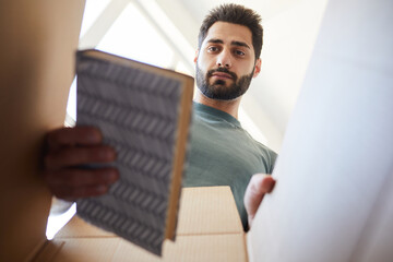 Young bearded man unpacking books from the box during his relocation to new house