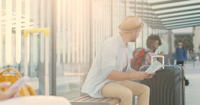 Young Caucasian handsome man traveller in hat sitting on bench on bus stop with city map and waiting for transport. Stylish male tourist looking for route on town plan.